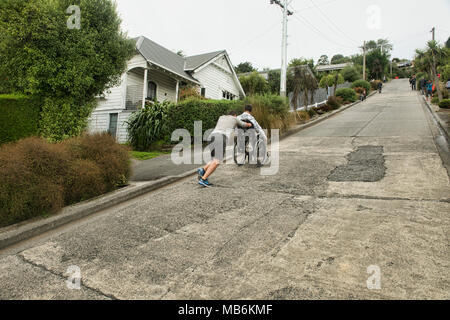 Freund seinen Kumpel Drücken in einem Rollstuhl bis Baldwin Street, der steilsten Strasse der Welt, Dunedin, Neuseeland Stockfoto