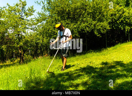 Nahaufnahme von Mann mähen Grün wildes Gras Feld mit freischneider Rasenmäher oder Power Tool string Rasentrimmer. Saisonale garten Reinigung und Mähen Konzept. Stockfoto