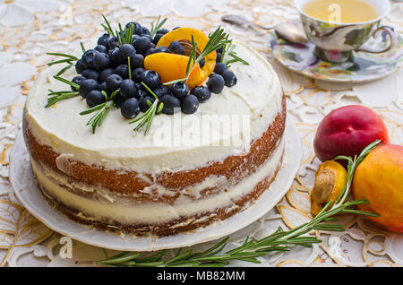 Seitenansicht des köstlichen hausgemachten Kuchen mit Sahne, Beeren, Rosmarin und Nektarine oben auf einem Dekorativen weißen Tischdecke neben einem vintage Tasse Tee Stockfoto