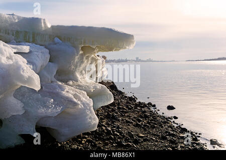 Schmelzenden Eisschollen auf dem steinigen Strand von einem breiten Flussbett vor dem Hintergrund einer unscharfen Stadt in der Ferne Stockfoto