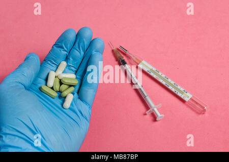Sortiment von Pillen, Tabletten und Kapseln auf Hand in Blau Handschuh gegen Rosa Hintergrund, Spritze und Thermometer. Stockfoto
