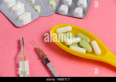 Sortiment von Pillen und Kapseln in Gelb Löffel, Spritze, Thermometer auf Rosa Hintergrund. Stockfoto