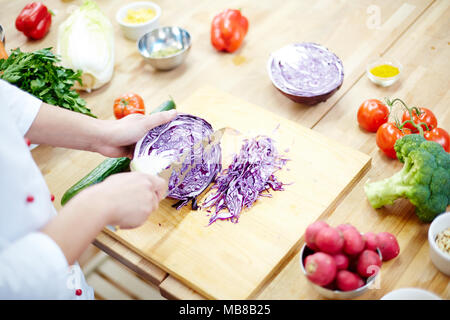 Hände von Küchenchef mit Messer schneiden frisches Kraut auf Holzbrett unter anderem frisches Gemüse im Prozess des Kochens Stockfoto