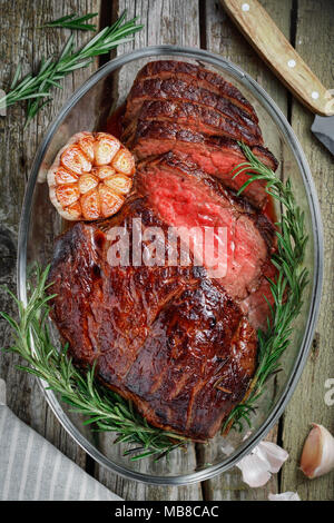 Blick von oben auf eine gebackene Rindfleisch Steak medium in Stücke schneiden in einer Glasschale mit gebackener Knoblauch und Rosmarin. Das Konzept einer festlichen Tisch und Küche. Stockfoto