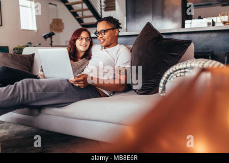 Mixed Race Paar sitzt auf einer Couch mit Laptop im Wohnzimmer. Paar entspannt auf einem Sofa mit Laptop zu Hause. Stockfoto