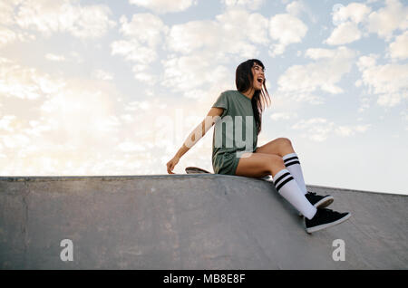 Lachend weiblichen Skateboarder sitzen auf einer Rampe im Skate Park. Frau sitzt auf der Skateboard- und Lachen. Stockfoto