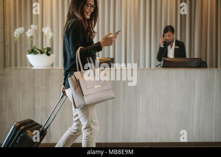 Frau mit Mobiltelefon und ihren Koffer ziehen in eine Hotellobby. Weibliche Business Reisenden zu Fuß im Hotel Flur. Stockfoto