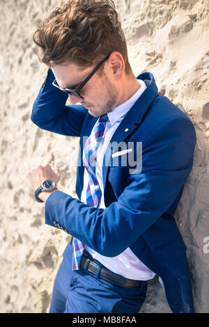 Ernsthaften jungen Mann im eleganten Anzug entspannt auf Sand. Stilvolle Kerl liegt am Strand und schaut auf die Armbanduhr. Lustig Manager in der Zeit verloren und wird spät Stockfoto