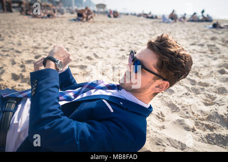 Ernsthaften jungen Mann im eleganten Anzug ist spät auf der Arbeit. Stilvolle Kerl liegt am Strand und sieht auf der Armbanduhr. Lustig Manager in der Zeit verloren Stockfoto