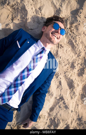 Ungewöhnliche junge Mann im eleganten Anzug entspannt am Strand und lächelnd. Lustige Tourist liegt auf den Sand und genießen Ferienhäuser Stockfoto