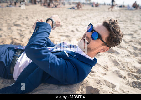 Ungewöhnliche junge Mann im eleganten Anzug entspannt am Strand und lacht. Lustiger Kerl legt sich auf den Sand und hat Zeit zum Ausruhen Stockfoto