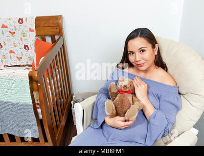 Attraktive schwangere Frau in einem Schaukelstuhl sitzend neben einem Kinderbett Holding einen Teddybären und lächelnd Stockfoto