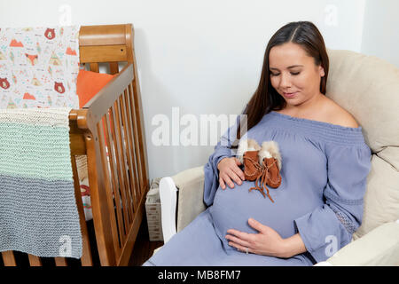 Attraktive schwangere Frau in einem Schaukelstuhl sitzt neben ein Kinderbett mit Baby Moccasins auf ihrem Bauch Stockfoto