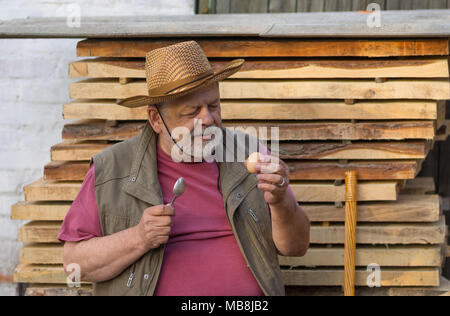 Bärtige senior fertig, rohe Hühnerei zu trinken, während im Freien sitzen gegen Stapel von Platten Stockfoto