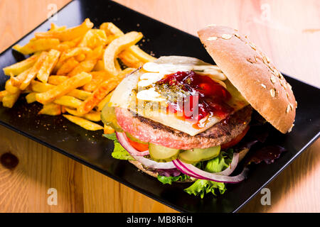 Hamburger mit Käse und Pickles auf schwarzem Teller serviert und begleitenden Kartoffeln Stockfoto