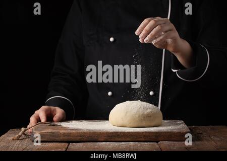 Koch Hände gießen Mehl Pulver auf rohem Teig mit Sieb auf schwarzem Hintergrund, Kochen Stockfoto