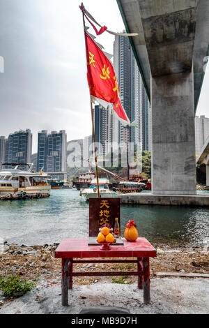 Eine rote Altar Tabelle enthält ein Angebot von Obst, Getränke und eine Schale mit Räucherstäbchen. Oben ist eine rote Flagge mit der chinesischen Schrift weht im Wind. Stockfoto