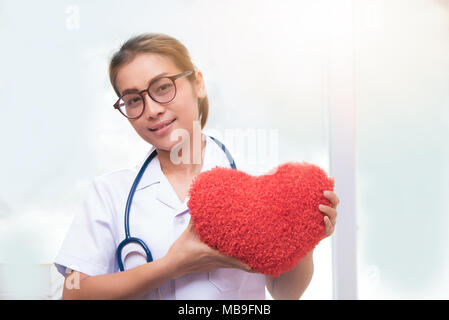 Frau Doktor halten roten Herzen, asiatische Arzt halten roten Herzen Kissen und blau Stethoskop halsschleife im Büro oder Arbeitszimmer Stockfoto
