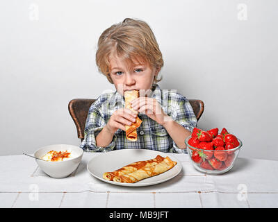 Süße Junge sitzt am Frühstückstisch essen Pfannkuchen und Erdbeeren Stockfoto