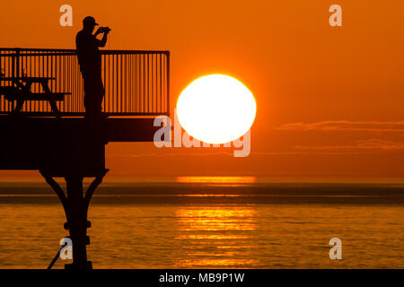 Aberystwyth Wales UK, Sonntag, 08. April 2018 UK Wetter: Ein Mann steht und Fotos am Ende abgeschnitten von Aberystwyth Pier am Meer, wie die Sonne schön über die Cardigan Bay am Ende einen warmen und sonnigen Frühling an der britischen Westküste Foto © Keith Morris/Alamy leben Nachrichten Stockfoto