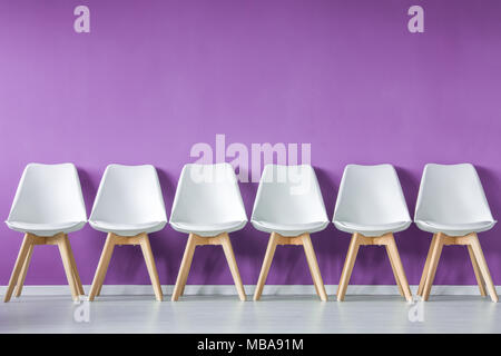 Vorderansicht aus einer Reihe von modern, einfach, weiß, Holz, leere Stühle gegen lila Wand in einem minimalistischen Interieur. Stockfoto