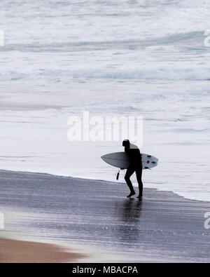 Die Silhouette einer erschöpften Surfer sein Surfbrett tragen als geht er aus dem Meer auf den Fistral Beach in Newquay Cornwall. Stockfoto
