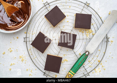 Caramel Shortbread/Millionäre shortbread Quadrate mit einem Palettenmesser und geschmolzene dunkle Schokolade in einer Schüssel auf einem weißen Hintergrund Stockfoto