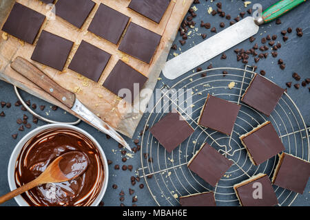 Caramel Shortbread/Millionäre shortbread Quadrate mit einem Palettenmesser und geschmolzene dunkle Schokolade in einer Schüssel auf einem Schiefer Hintergrund Stockfoto