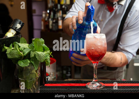 Nahaufnahme von einem Barkeeper mit einem roten Papillon, aus einem rotem spritz auf eine Bar couter. Er giesst Soda Wasser in das Glas voller Eis. Stockfoto