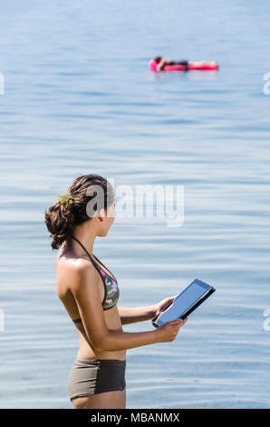 Porträt der jungen Frauen, die in den Bodensee mit digitalen Tablet in der Hand und eine andere Person auf der Luftmatratze im Hintergrund Stockfoto