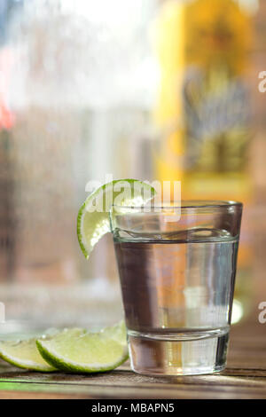 Alkohol shot Glas, Tequila oder Wodka, mit einem Kalk/Zitronenscheibe, Nahaufnahme, mit vielen Flaschen im Hintergrund, unscharf Stockfoto