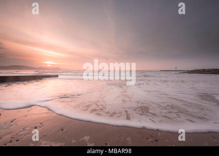 Schöne rosa Töne in den Himmel und Meer bei Sonnenaufgang als morgen Wellen sanft Rolle auf dem Sandstrand in Lyme Regis. Stockfoto