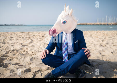 Junge trendige Mann in lustige Maske und eleganten Anzug auf Urlaub. Einhorn sitzt am Strand auf dem Hintergrund von Himmel und Meer Stockfoto