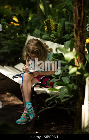 Ein zehn Jahre altes Mädchen sitzen auf einer Bank, Zeichnung Blumen in einem Gewächshaus. Stockfoto
