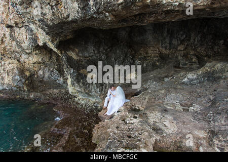 Paar Brautpaar Braut und Bräutigam Lachen und Lächeln zu einander, glücklichen und freudigen Moment. Der Mann und die Frau in der Hochzeit Kleidung sitzen auf dem Rock Hintergrund. Stockfoto