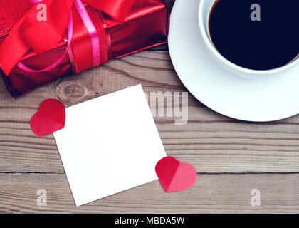 Roter Geschenkbox mit Schleife auf einer hölzernen Hintergrund. Eine Tasse Kaffee und einen gestrickten Herz. Valentinstag. Stockfoto