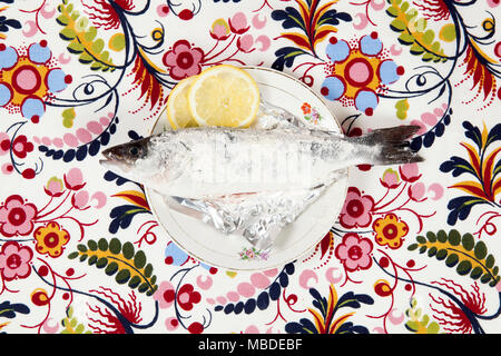 Eine Zubereitung aus einem Baß-Fischen mit einer Scheibe Zitrone neben innerhalb einer Blume Platte auf einem geblümten Stoff versteckt. Camouflage Spiel. Minimale Farbe noch Leben Stockfoto