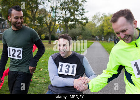 Lächelnd männliche Läufer Händeschütteln mit Freund im Rollstuhl an der Nächstenliebe Rennen in Park Stockfoto