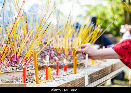 Nahaufnahme der Räucherstäbchen und Kerzen in Sand auf einem Tisch gepflanzt während einer buddhistischen Zeremonie, mit weiblicher Hand im Hintergrund. Stockfoto