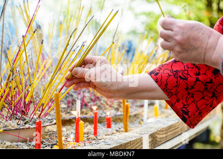 Nahaufnahme der Hände einer alten Frau das Tragen eines asiatischen Stil Bluse kleben rauchen Räucherstäbchen in ein Sand während einer buddhistischen Zeremonie. Stockfoto