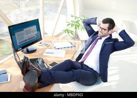 Die volle Länge der erfolgreiche junge Unternehmer tragen Anzug mit seinem modernen Büro sitzt mit seinen Beinen auf Büro Schreibtisch. Stockfoto
