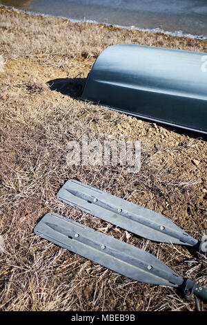 Zwei Ruder mit Aluminium Rotorblätter auf dem Boden liegend mit getrocknetem Gras entlang eines kleinen umgeworfen Beiboot auf einer Bank über Wasser Stockfoto