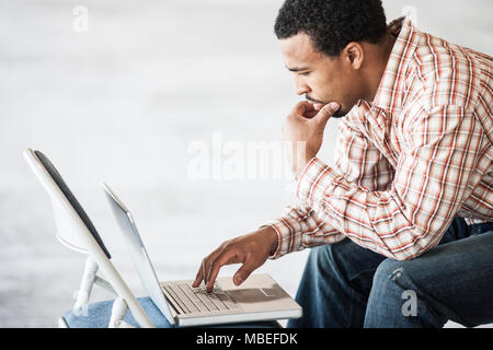 Schwarzer Mann bei der Arbeit auf einem Laptop und usiing einen Stuhl auf einen Tisch. Stockfoto