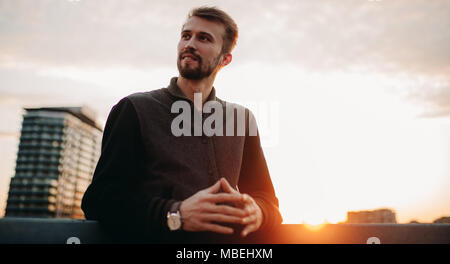 Glückliche junge Mann steht und lächelt auf Haus Dach vor Hintergrund der Wolkenkratzer und den Sonnenuntergang. Nahaufnahme. Kopieren Sie Platz. Stockfoto