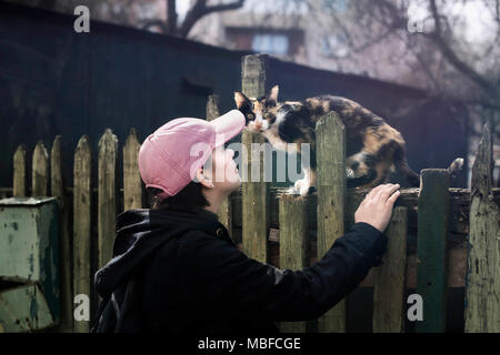 Ein Mädchen in einem rosa Baseball Cap und eine schwarze Jacke neben eine streunende Katze auf einem alten grünen Zaun. Minsk, Weißrussland. Stockfoto