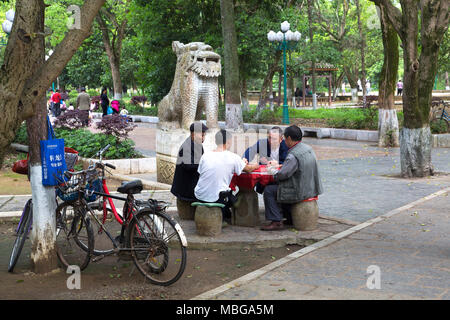 Vier chinesische Männer sitzen, die Karten in Yangshuo, China. Ihre Fahrräder gegen einen Baum. Hinter einem Stein Chinesischen guardian Löwe steht. Stockfoto