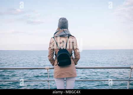 Rückansicht der jungen Frau in beige Mantel, Schal, Hut und Rucksack am Meer. Weibliche Reisende hält ihre Hände in den Taschen steht am Kai. Stockfoto