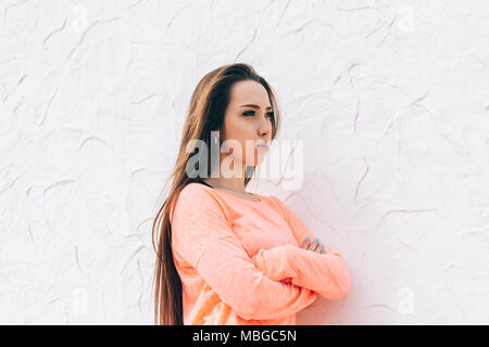 Beleidigt, schöne junge Frau, die in der Nähe von weißen Wand und weg schauen mit verschränkten Armen. Portrait von unglücklichen Frau mit langen braunen Haaren fühlt sich Angr Stockfoto