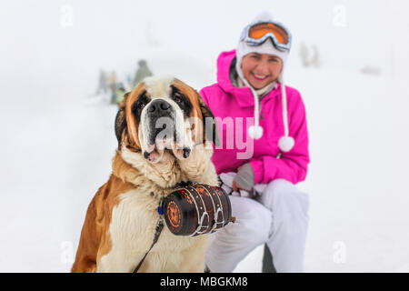 Bernhardiner Hund mit iconic Fass sitzt im Schnee mit unscharfen junge Frau in rosa Winterjacke im Hintergrund. Stockfoto