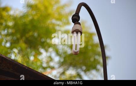 Beschädigte energy saver Glühbirne hängen mit einem korrodierte Lamp Post in der Öffentlichkeit mit Baum im Hintergrund Stockfoto